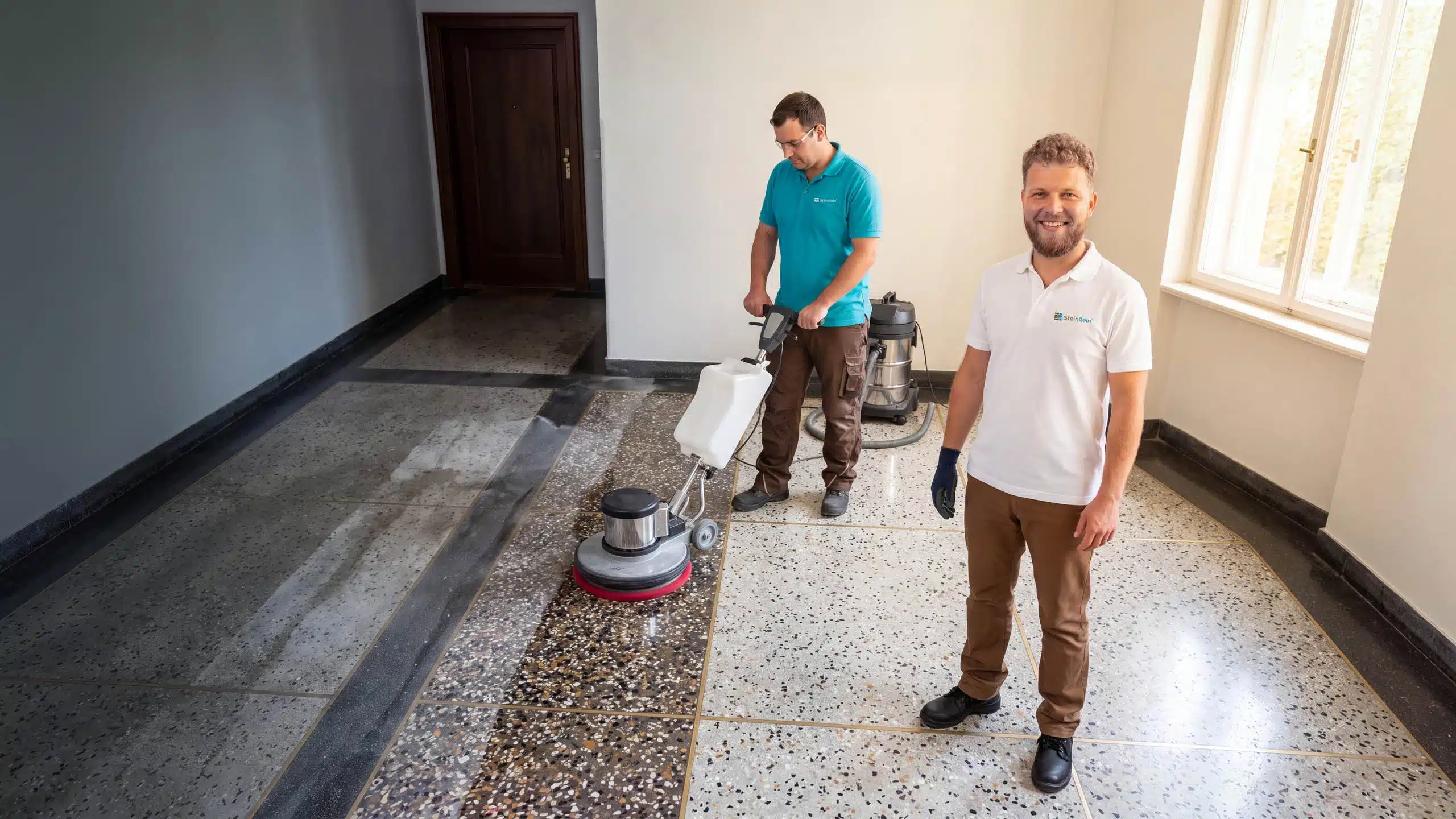 Symbolbild: Terrazzo reinigen – ein SteinRein-Techniker reinigt einen verschmutzten Terrazzoboden im Altbau-Hausflur mit der Einscheibenmaschine, der frisch gereinigte Streifen glänzt nass, vorn ein Kundenberater im weißen SteinRein-Polo.
